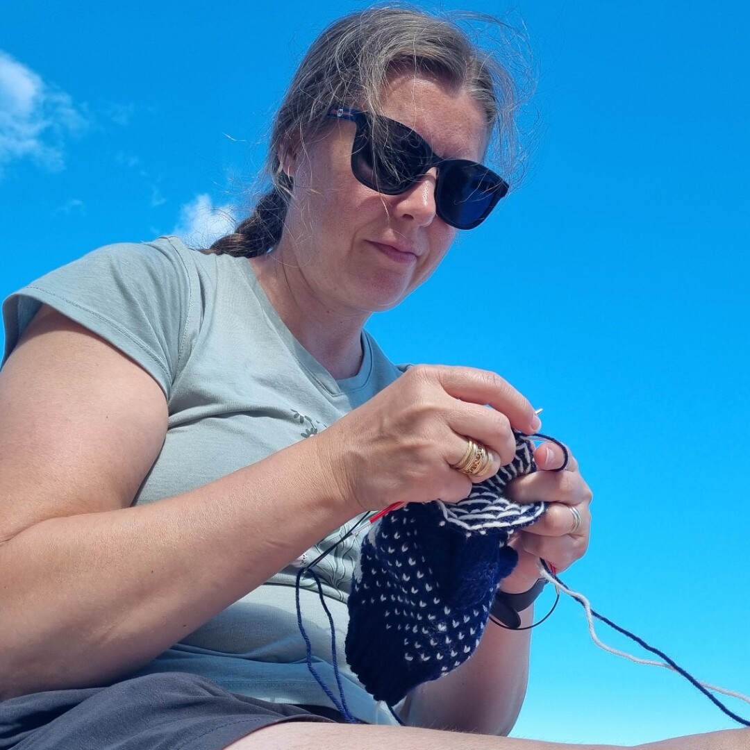 Aurinkolasit ja vihreän paidan pukeutunut nainen istuu ulkona aurinkoisena päivänä ja neuloo mustalla ja valkoisella langalla kirkkaan sinistä taivasta ja siellä täällä leijuvia pilviä vasten.