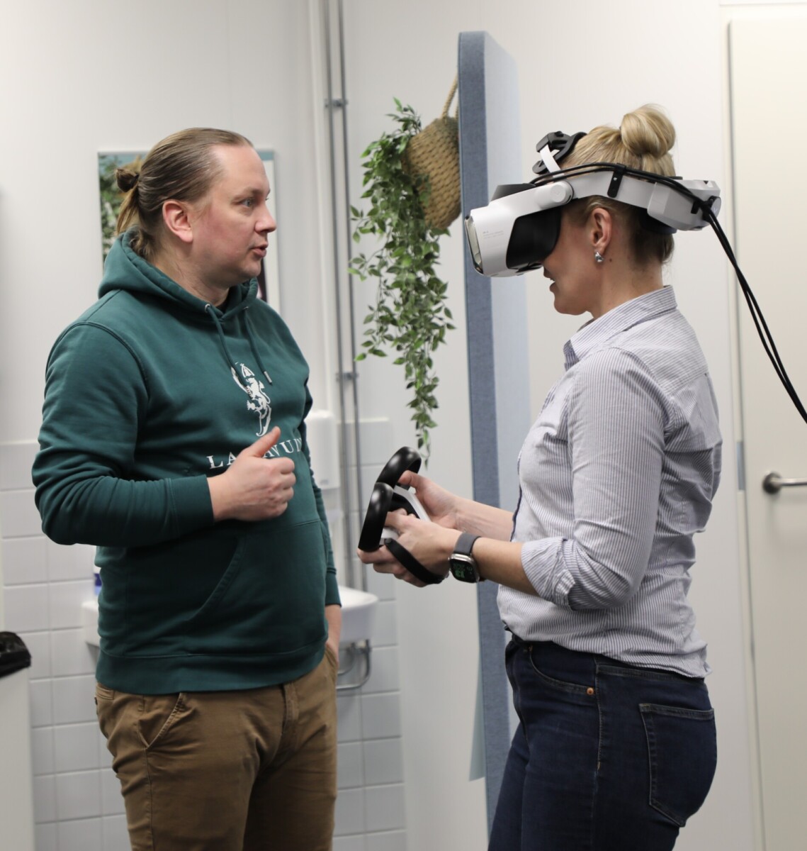 Vihreää hupparia pukeutunut mies keskustelee naisen kanssa, jolla on päässään VR-lasit ja kädessään ohjaimet. He ovat modernissa sisätilassa, jonka seinät ovat valkoiset ja taustalla on kasvi.