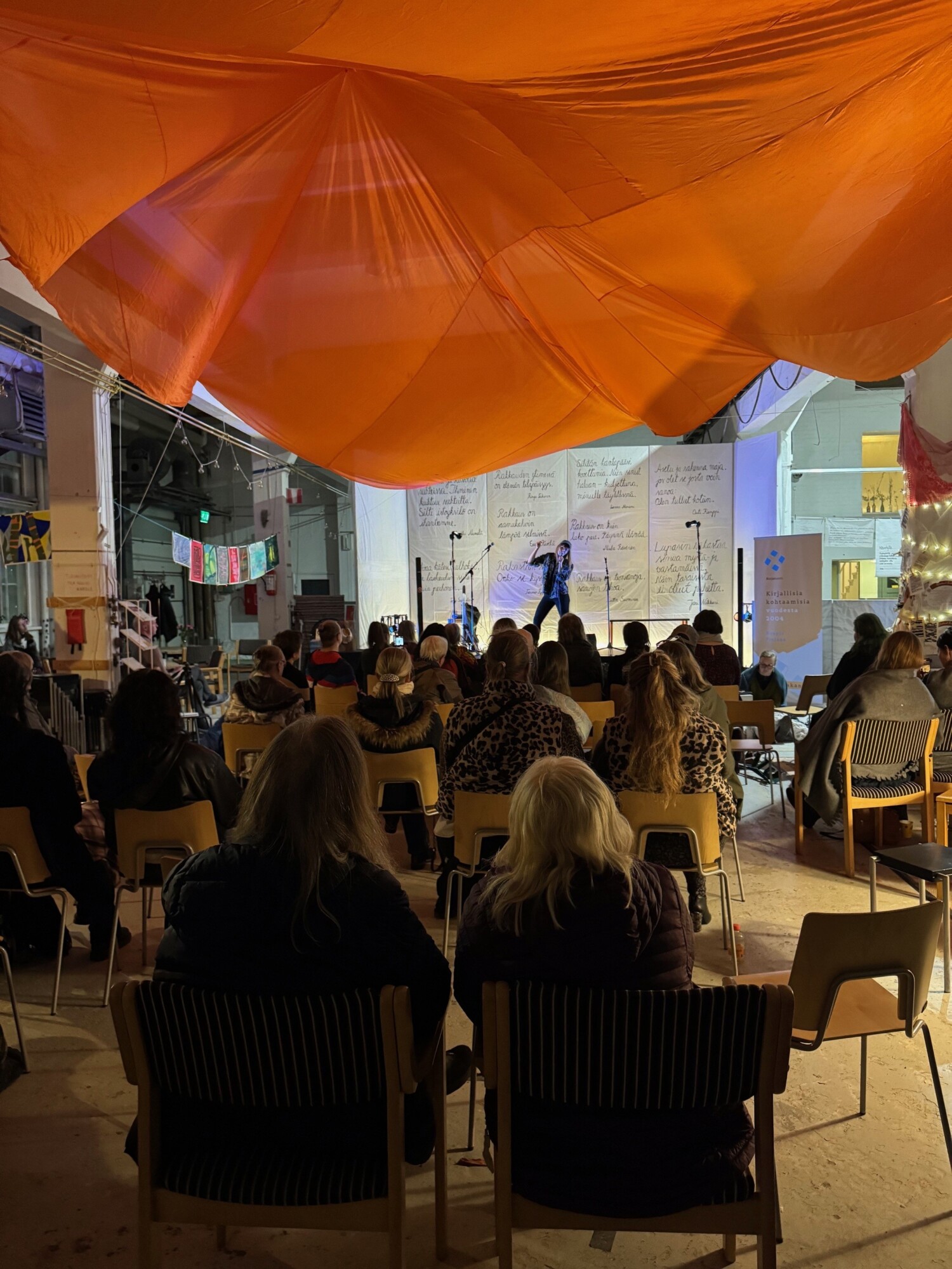 Yleisö istuu sisätiloissa ja seuraa esiintyjää lavalla suuren oranssinvärisen katoksen alla; lavan takaseinällä on käsinkirjoitettuja julisteita ja sisätiloissa on pehmeä valaistus.