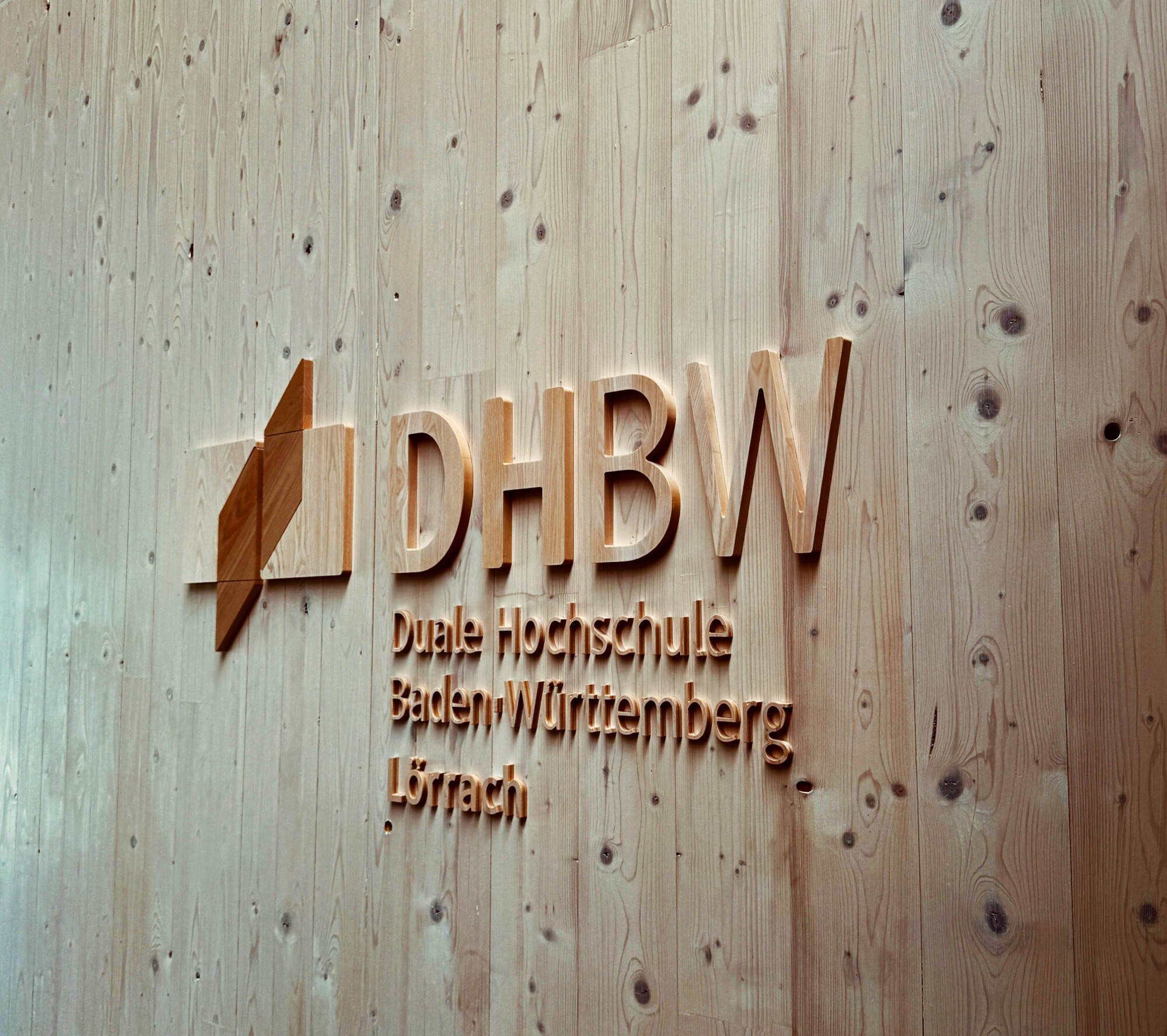 Puinen seinä, jossa on kohokuvioidut kirjaimet, jotka esittävät logon sekä nimet ”DHBW” ja ”Duale Hochschule Baden-Württemberg Lörrach” saksaksi. Teksti ja logo ovat modernia, minimalistista tyyliä.