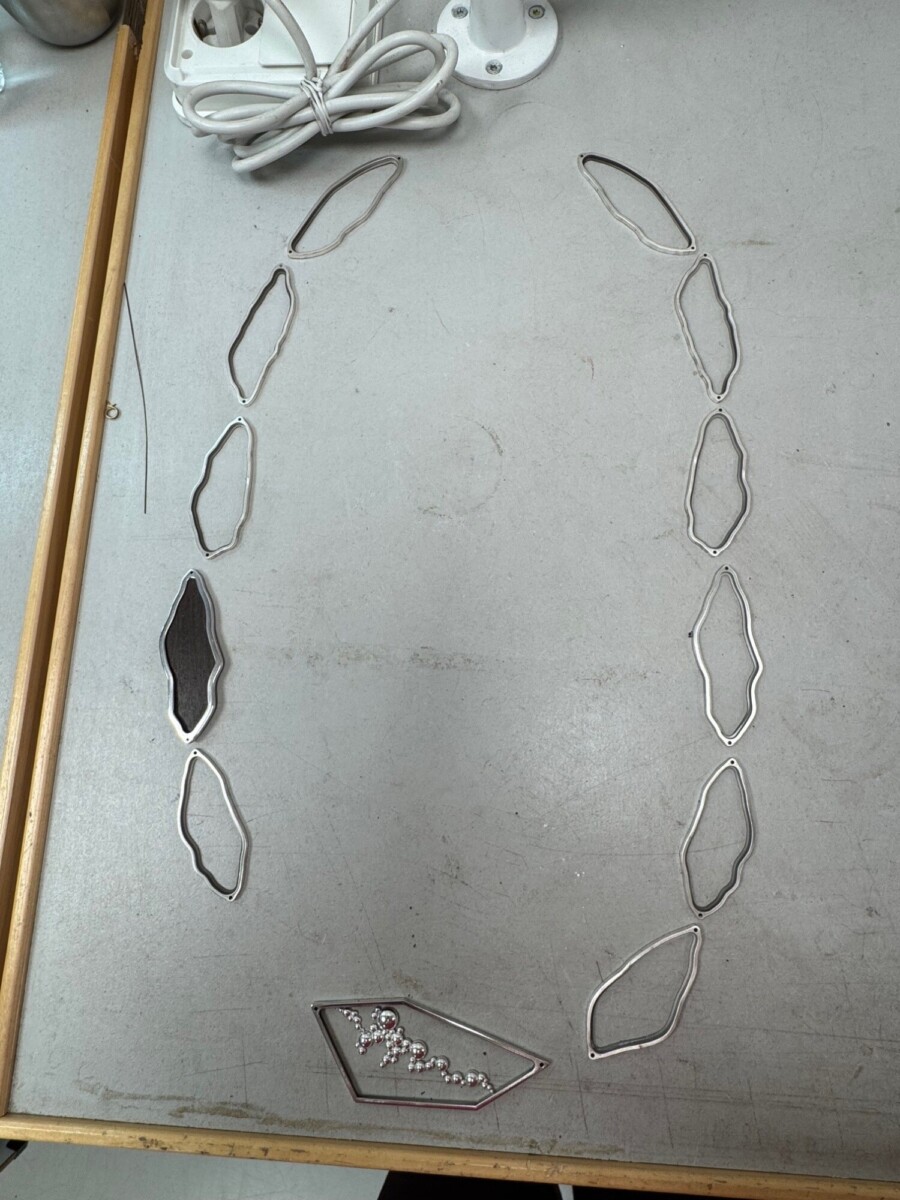 Kaksitoista epäsäännöllisen muotoista hopeanväristä metallikappaletta ja yksi pieniä helmiä sisältävä kappale on aseteltu harmaalle pöydälle valkoisen narun viereen.