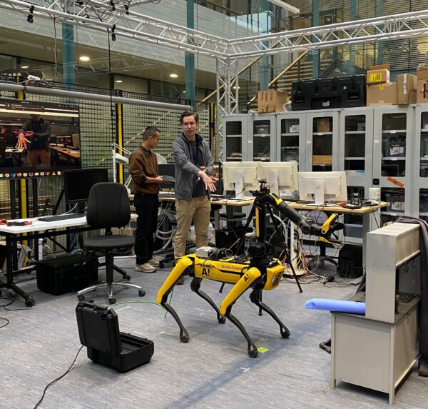 Kaksi ihmistä seisoo laboratoriossa, jossa on tietokoneita ja laitteita, ja vuorovaikuttaa keltaisen robottikoiran kanssa. Robottikoira on keskellä, ja sitä ympäröivät työpöydät, näytöt, tuolit ja johdot modernissa työtilassa.