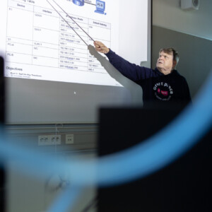 Tumman hupparin pukeutunut henkilö osoittaa luokkahuoneen valkoiselle valkokankaalle heijastettua kaaviota; etualalla näkyy tietokoneiden näyttöjä ja kaapeleita.