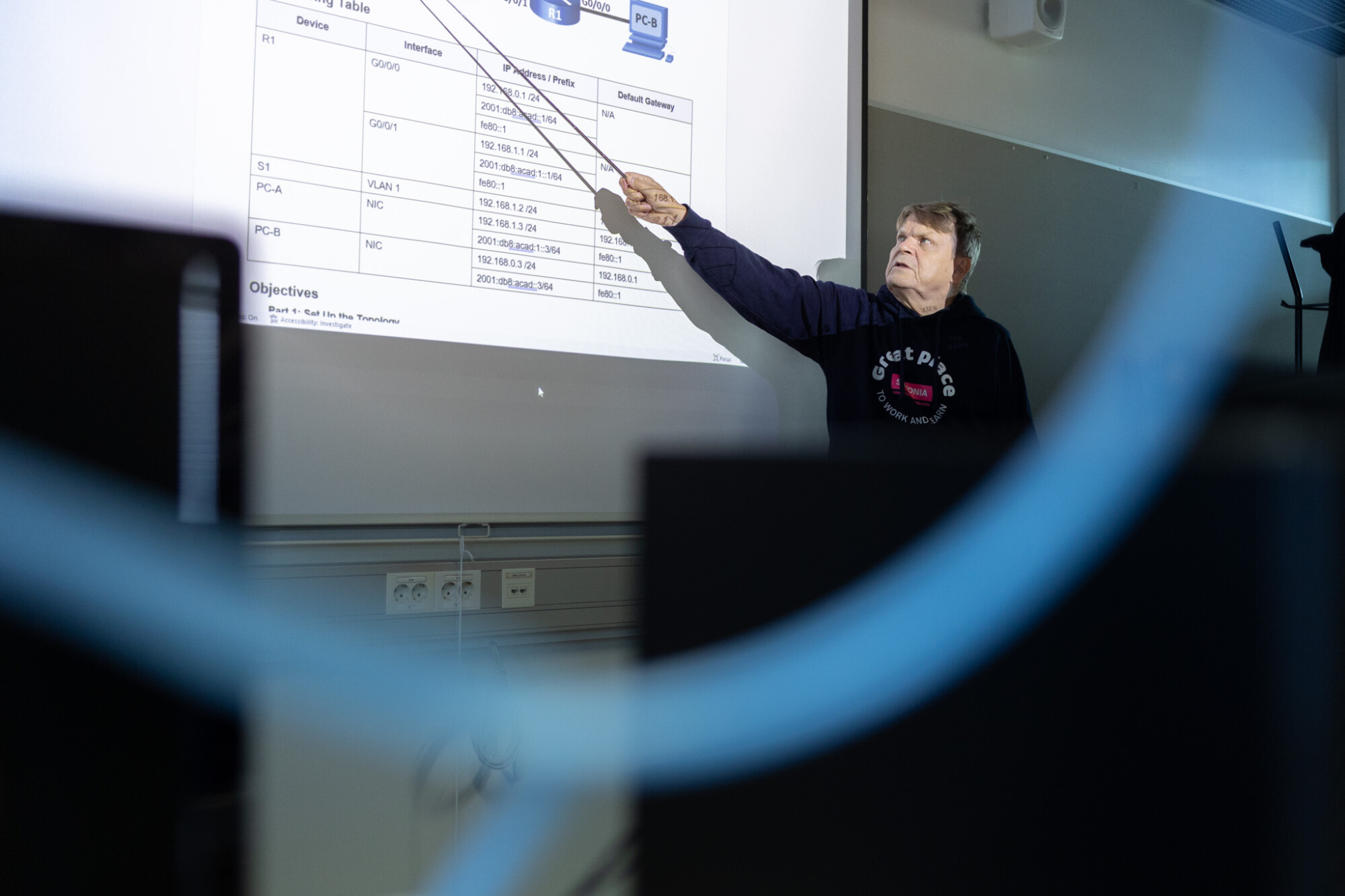 Tumman hupparin pukeutunut henkilö osoittaa luokkahuoneen valkoiselle valkokankaalle heijastettua kaaviota; etualalla näkyy tietokoneiden näyttöjä ja kaapeleita.