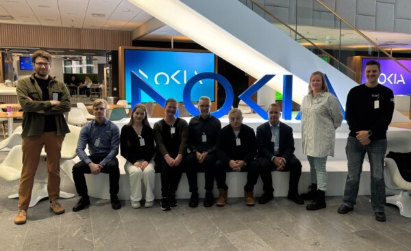 Yhdeksän ihmistä poseeraa sisätiloissa suuren valaistun Nokia-kyltin edessä, osa istuen ja osa seisoen, modernissa toimistotilassa, jonka taustalla näkyy portaikko ja näyttöjä.