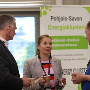 Kolme henkilöä seisoo ja keskustelee kahvikupit kädessään verkostoitumistilaisuudessa. Heidän takanaan on suomenkielinen kyltti, jossa lukee ”Pohjois-Savon energiaklasteri”. He näyttävät olevan keskittyneitä keskusteluun konferenssitilaisuudessa.