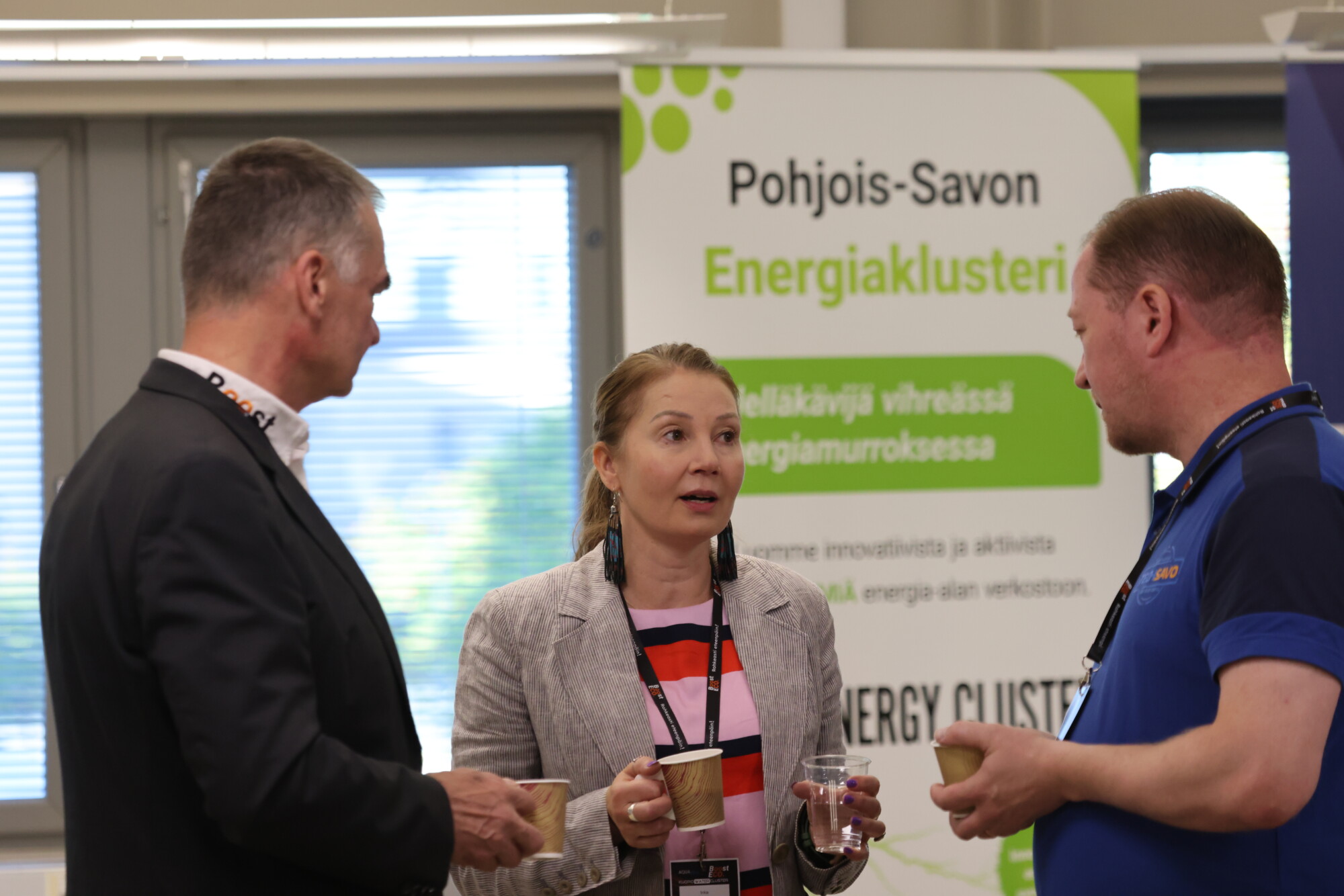 Kolme henkilöä seisoo ja keskustelee kahvikupit kädessään verkostoitumistilaisuudessa. Heidän takanaan on suomenkielinen kyltti, jossa lukee ”Pohjois-Savon energiaklasteri”. He näyttävät olevan keskittyneitä keskusteluun konferenssitilaisuudessa.