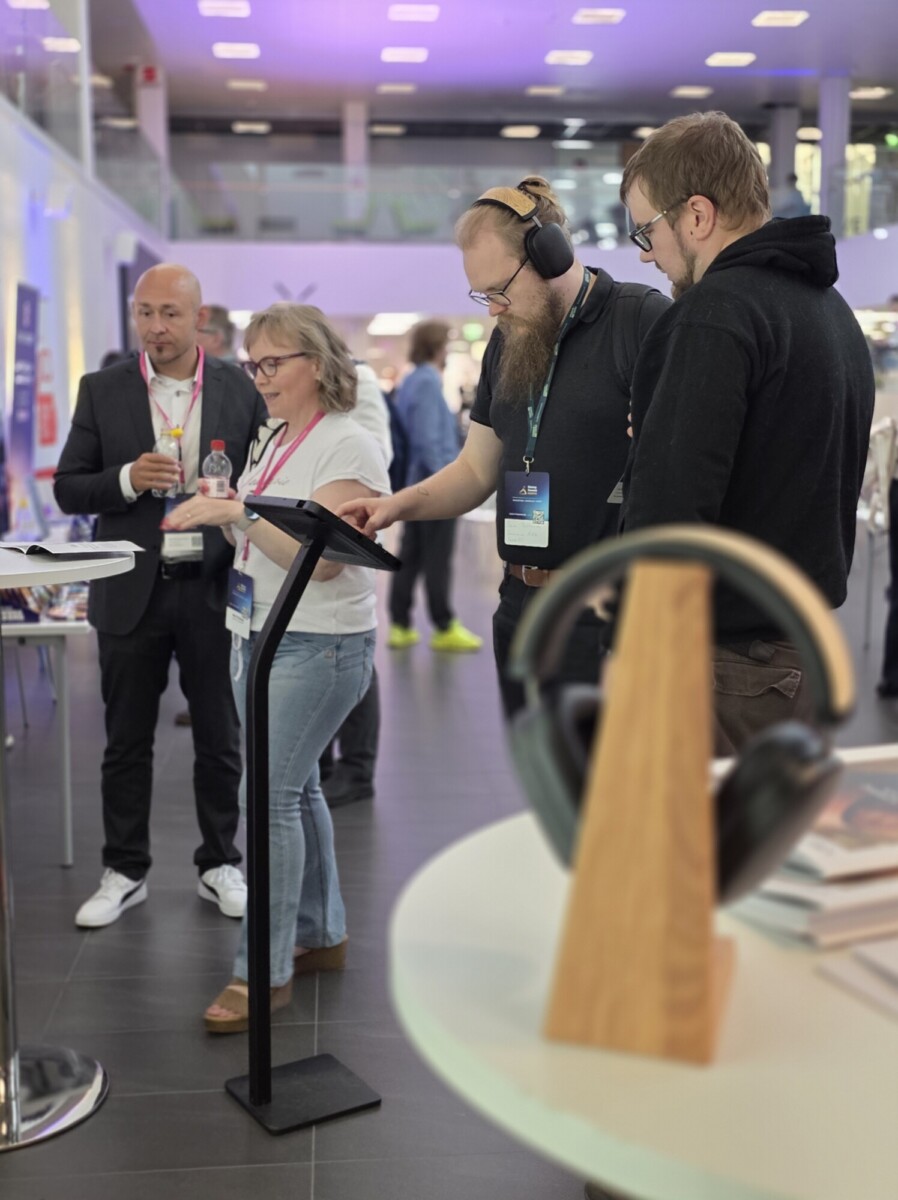 Neljä henkilöä seisoo ja keskustelee sisätiloissa järjestettävässä tapahtumassa tai konferenssissa. Kaksi miestä käyttää kuulokkeita ja kosketusnäyttöä, kun taas nainen ja mies keskustelevat lähellä. Epäterävässä etualassa näkyy kuulokkeet ja esillä olevia kirjoja.