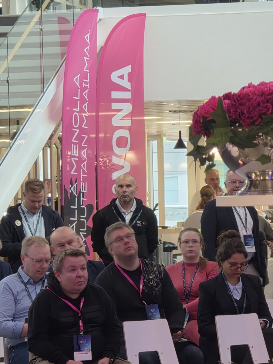 Ryhmä ihmisiä istuu ja kuuntelee sisätiloissa järjestettävässä tilaisuudessa, jonka taustalla näkyy kaksi korkeaa vaaleanpunaista lippua ja suuri vaaleanpunaisista kukista koostuva maljakko.