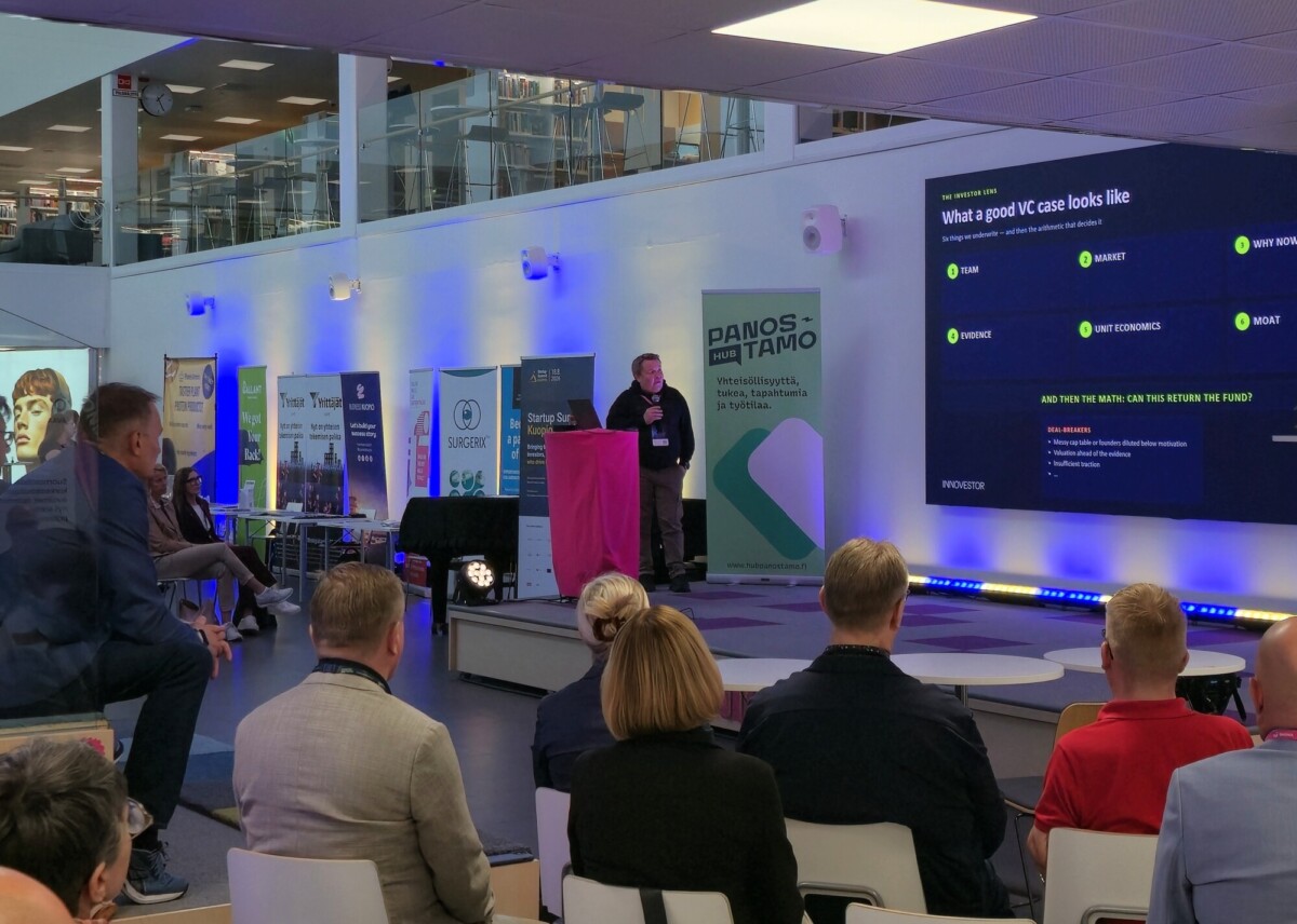 Puhuja pitää esitelmää istuvan yleisön edessä modernissa, hyvin valaistussa kokoustilassa, jossa on bannereita ja suuri näyttö, jolla näkyy otsikko ”Miltä hyvä VC-tapaus näyttää…”.