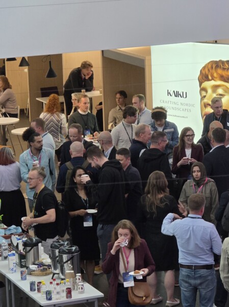 Ihmisjoukko seurustelee, syö ja juo konferenssitilaisuudessa. Osa seisoo ryhmissä, toiset istuvat pöydän ääressä. Taustalla näkyy kyltti, jossa lukee ”Kaiku Crafting Nordic Soundscapes”.