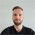 Mies, jolla on lyhyet vaaleanruskeat hiukset ja parta ja joka on pukeutunut mustaan paitaan, seisoo vaaleanvärisen sileän seinän edessä ja katsoo suoraan kameraan ilmeettömästi.