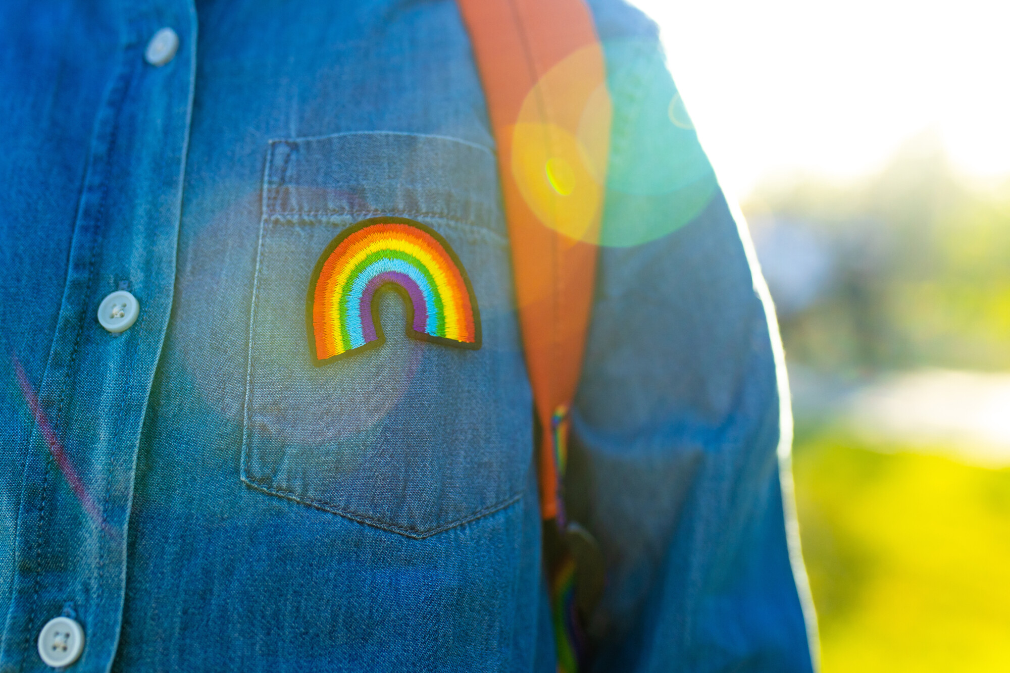 Henkilö, jolla on yllään denimpaita, jonka taskussa on sateenkaarimerkki, sekä reppu, seisoo ulkona kirkkaassa auringonvalossa, ja kuvassa näkyy linssiheijastuma.