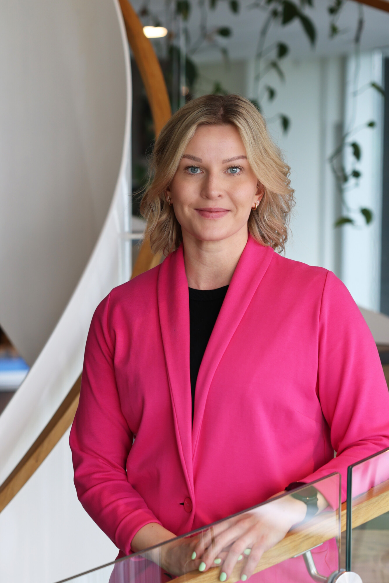 Vaaleatukkainen nainen, jolla on yllään kirkkaan vaaleanpunainen bleiseri ja musta paita, seisoo sisätiloissa ja hymyilee kameraan. Taustalla näkyy kasveja ja kierreportaat.