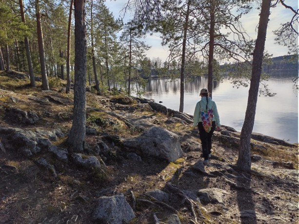 Henkilö, jolla on yllään kevyt takki ja värikäs laukku, kävelee kallioisen ja metsäisen alueen läpi rauhallisen järven rannalla osittain pilvisellä säällä. Korkeat puut ympäröivät maisemaa.