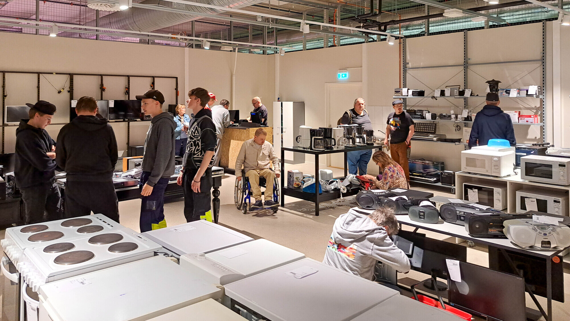 Ihmiset työskentelevät ja ovat vuorovaikutuksessa hyvin valaistussa elektroniikkapajassa, joka on täynnä laitteita, kuten mikroaaltouuneja, näyttöjä ja liesiä. Hyllyillä ja pöydillä on järjestyksessä erilaisia elektroniikkalaitteita ja komponentteja.