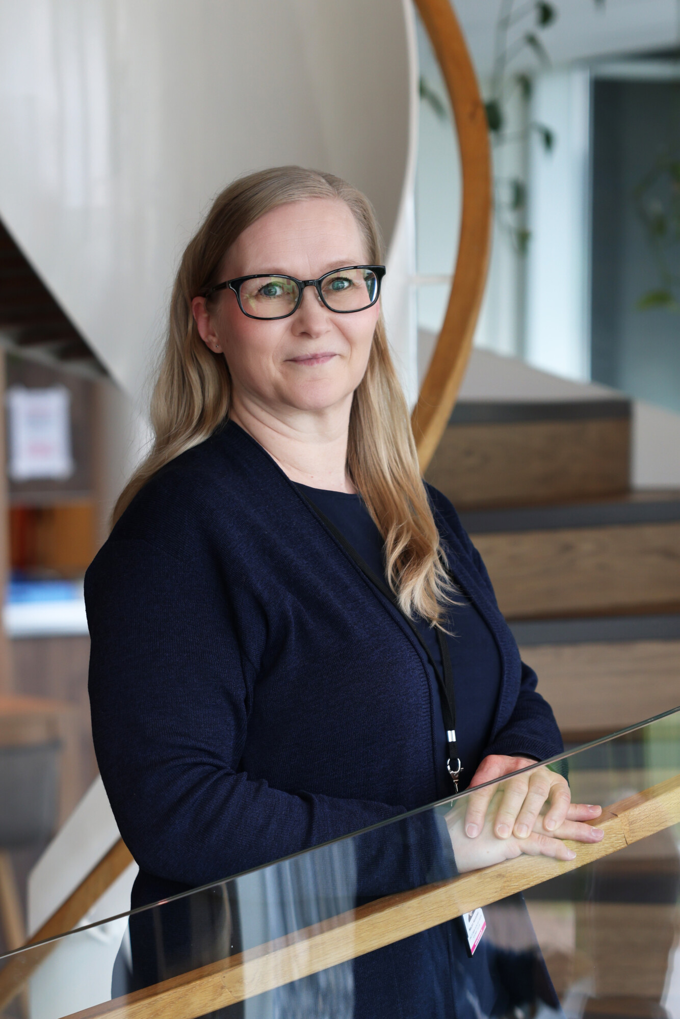 Nainen, jolla on pitkät vaaleat hiukset ja silmälasit ja joka on pukeutunut tummansiniseen neuletakkiin, seisoo sisätiloissa portaikon vieressä, kädet lasikaiteella, ja hymyilee hieman kameraan.