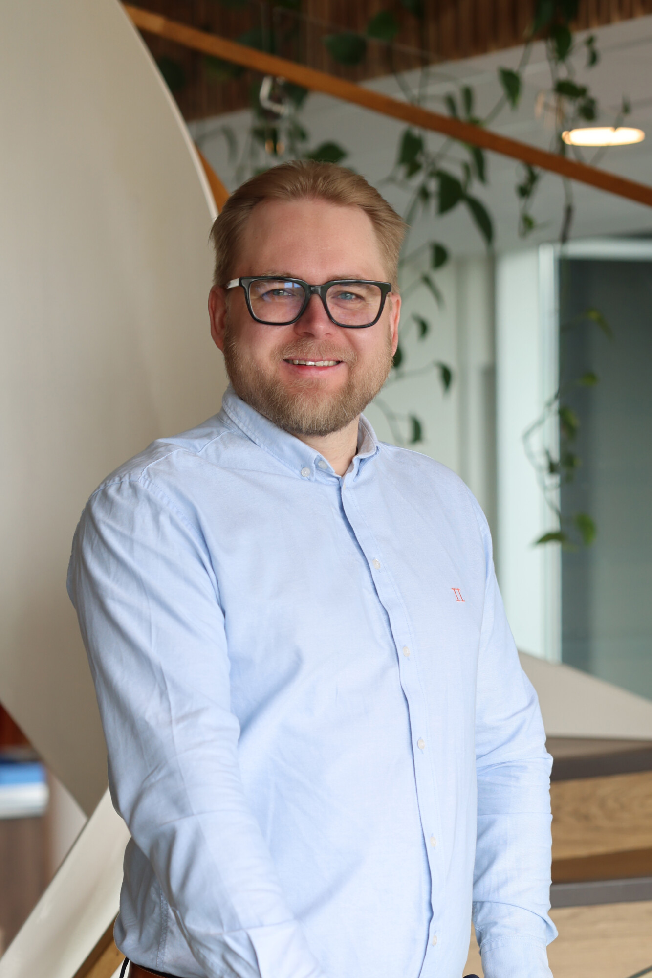 Hymyilevä mies, jolla on silmälasit ja vaaleansininen napituspaita, seisoo sisätiloissa modernin taustan edessä, jossa on kasveja ja puisia yksityiskohtia.
