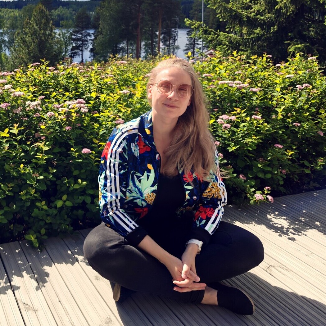 Nainen, jolla on pitkät vaaleat hiukset, silmälasit ja värikäs takki, istuu ristikkäin puuterassilla, ympärillään vihreitä, vaaleanpunaisilla kukilla koristeltuja pensaita, ja taustalla näkyy puita ja järvi.