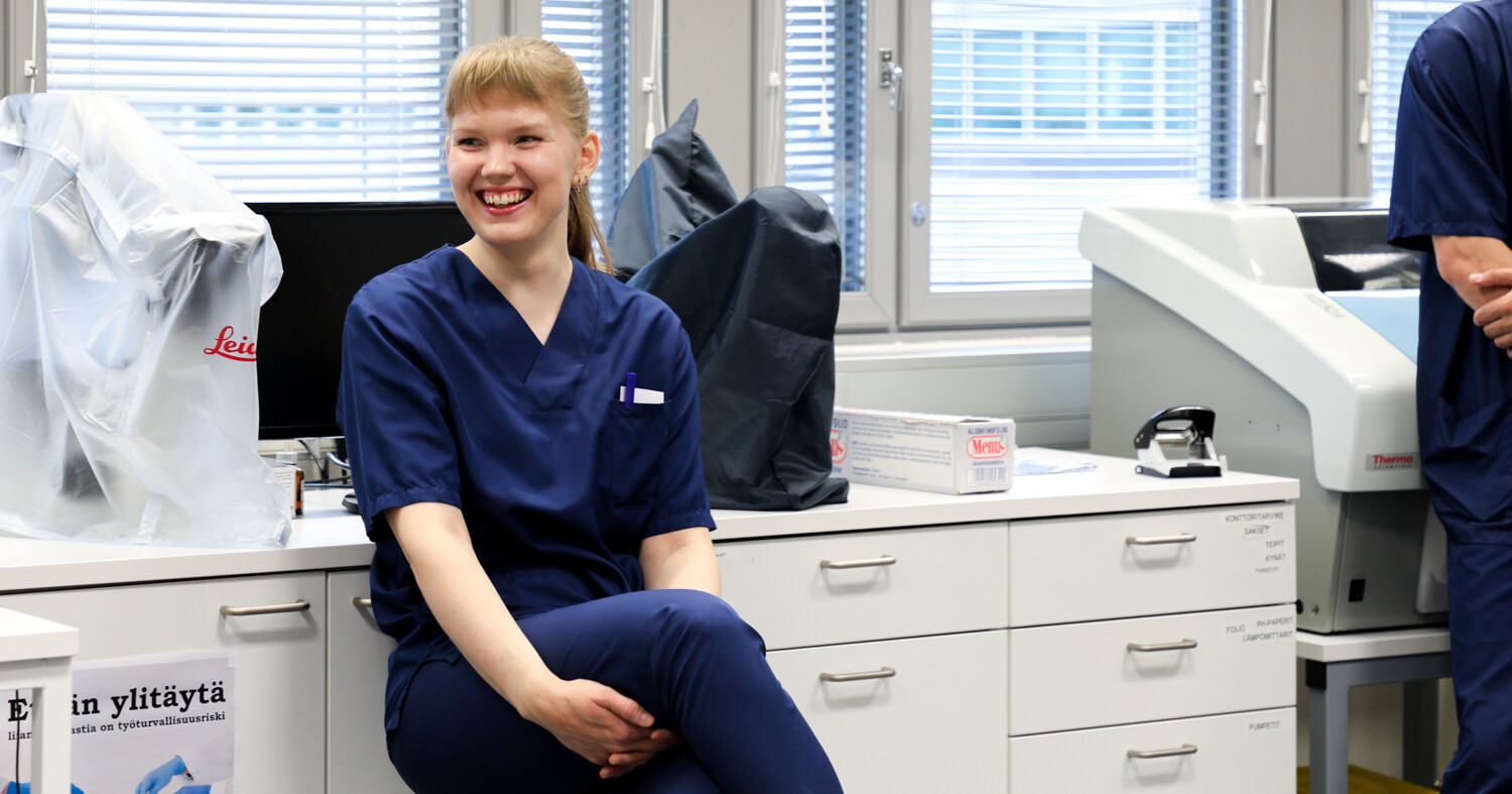 Sinisissä leikkaussalivaatteissa oleva henkilö istuu hymyillen lääketieteellisen laboratorion työtasolla. Taustalla näkyy tietokoneita, laitteita sekä toinen henkilö, joka näkyy osittain oikealla.