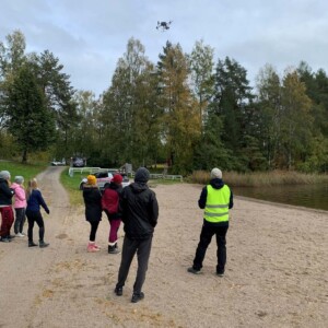 Ryhmä ihmisiä seisoo hiekkarannalla ja katselee yläpuolella lentävää dronea. Joillakin on päällään takit ja hatut, mikä viittaa viileään säähän. Taustalla näkyy puita ja polku.
