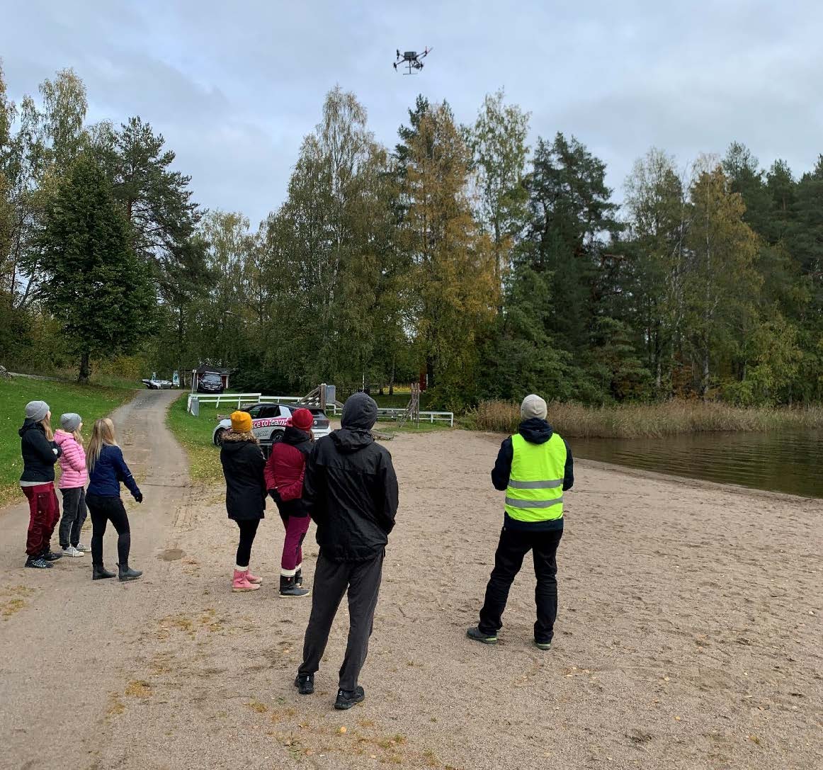 Ryhmä ihmisiä seisoo hiekkarannalla ja katselee yläpuolella lentävää dronea. Joillakin on päällään takit ja hatut, mikä viittaa viileään säähän. Taustalla näkyy puita ja polku.