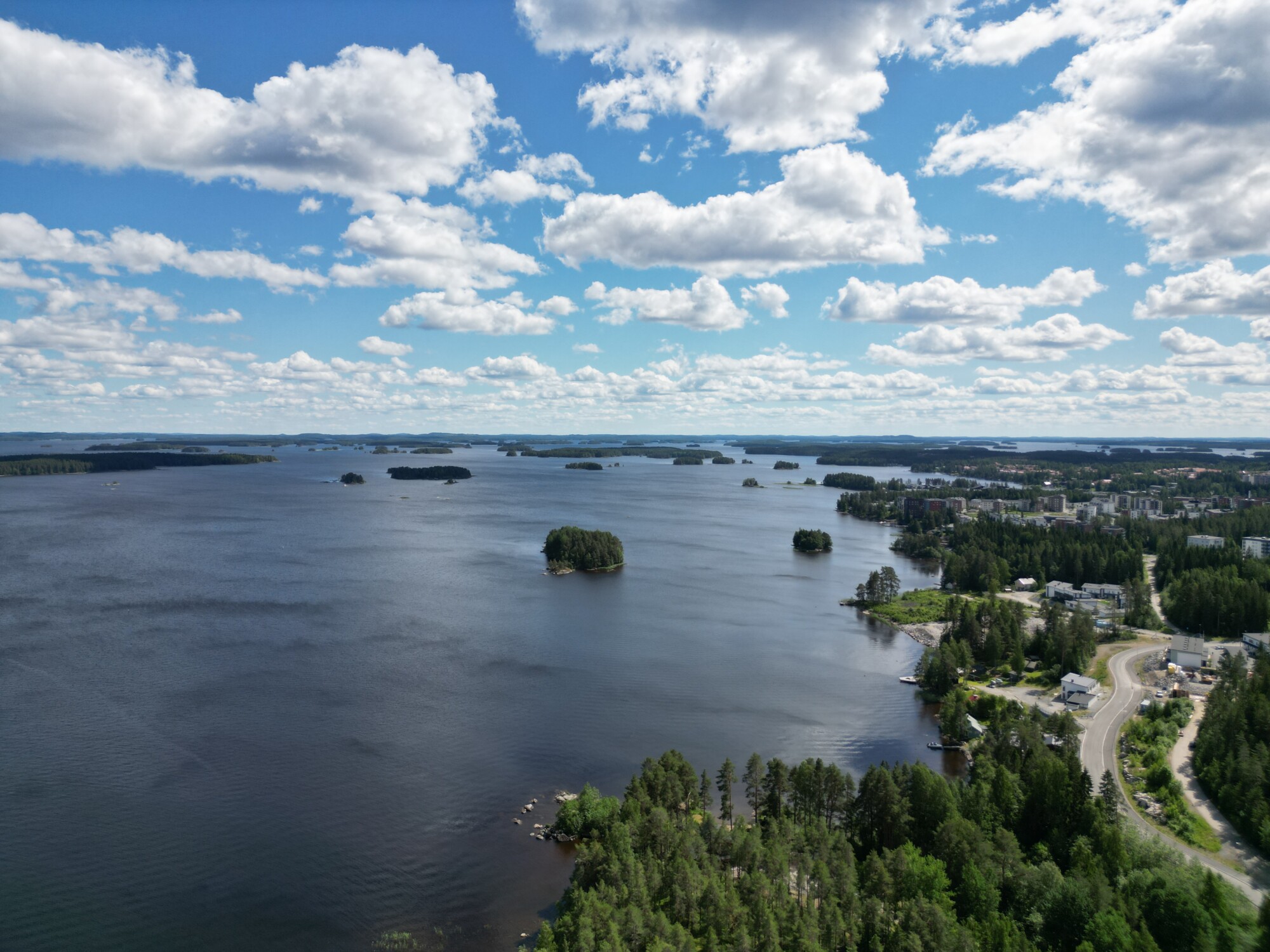Ilmakuva järvestä, jossa on pieniä metsäisiä saaria ja jota ympäröivät puut sekä muutama rakennus osittain pilvisen sinisen taivaan alla.