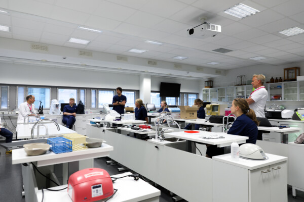 Ryhmä laboratoriotakkeihin ja leikkaussalivaatteisiin pukeutuneita ihmisiä istuu ja seisoo valkoisten pöytien ympärillä modernissa laboratoriossa keskustellen. Työtasoilla näkyy laboratoriolaitteita ja -tarvikkeita.
