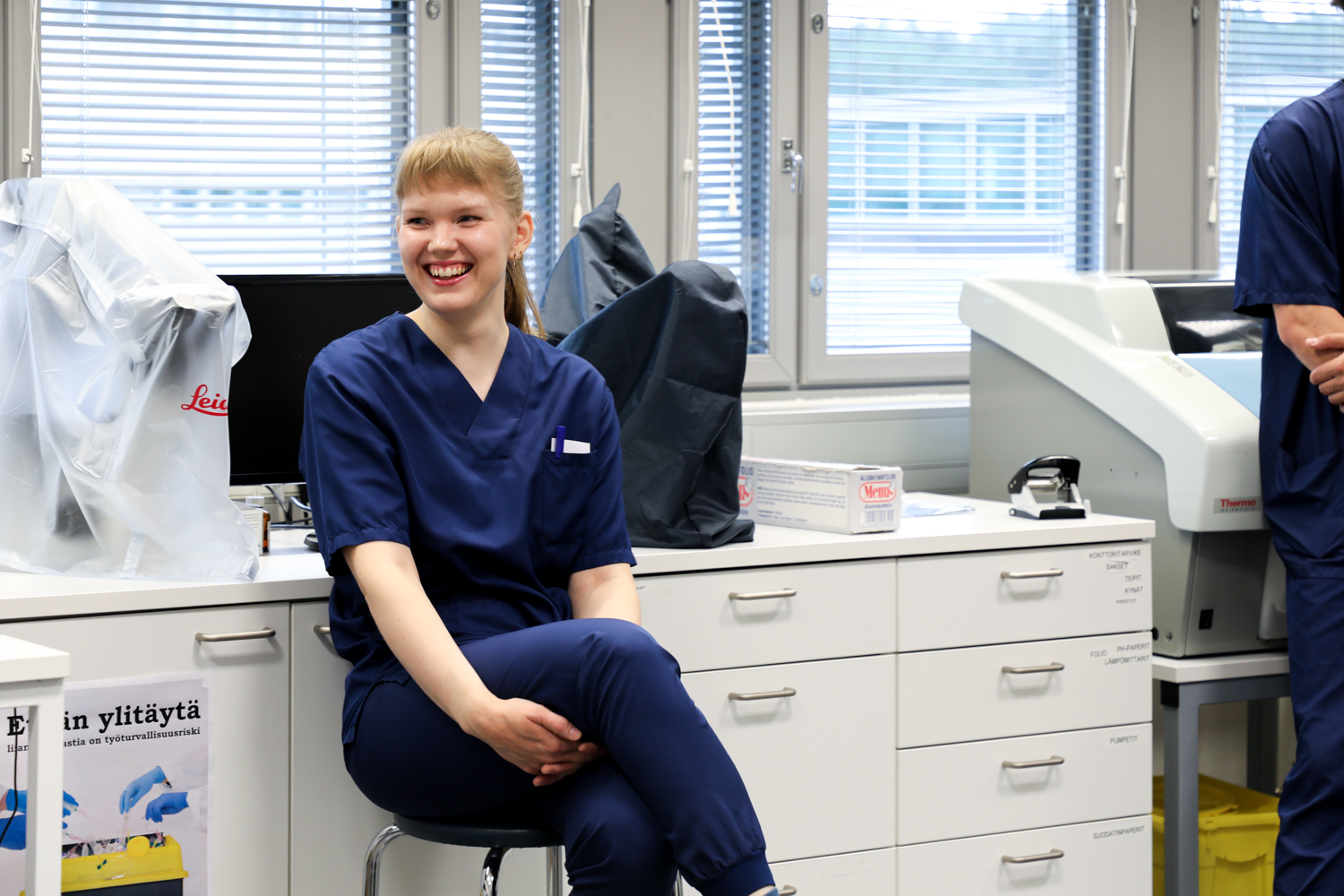 Sinisissä leikkaussalivaatteissa oleva henkilö istuu jakkaralla hymyillen valoisassa sairaala- tai laboratoriotilassa, jonka taustalla näkyy laitteita ja kaappeja.