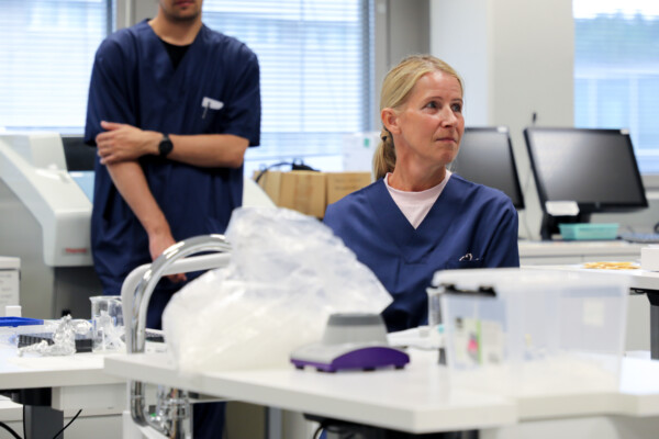 Kaksi henkilöä, joilla on yllään tummansiniset leikkaussalivaatteet, on valoisassa laboratoriossa, jossa on laitteita ja tietokoneita. Toinen henkilö istuu ja katsoo oikealle, kun taas toinen seisoo kädet ristissä, pääosin kuvan ulkopuolella. Läheisellä pöydällä on laboratoriotarvikkeita.