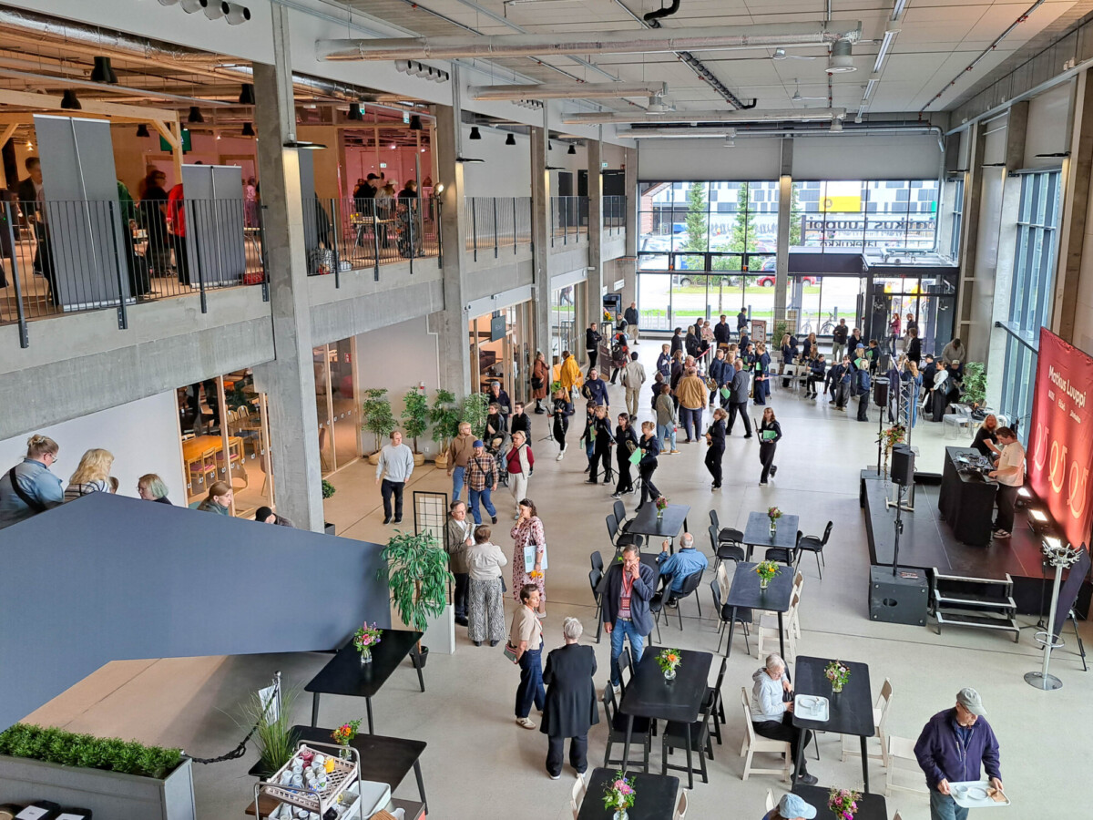 Suuri avoin sali, jossa ihmiset seurustelevat; osa istuu pöydissä, osa seisoo. Toisessa kerroksessa on parveke, jolla on lisää ihmisiä, suuret ikkunat sekä oikealla puolella punaisella taustalla oleva baaritiski. Tila vaikuttaa modernilta ja kutsuvalta.