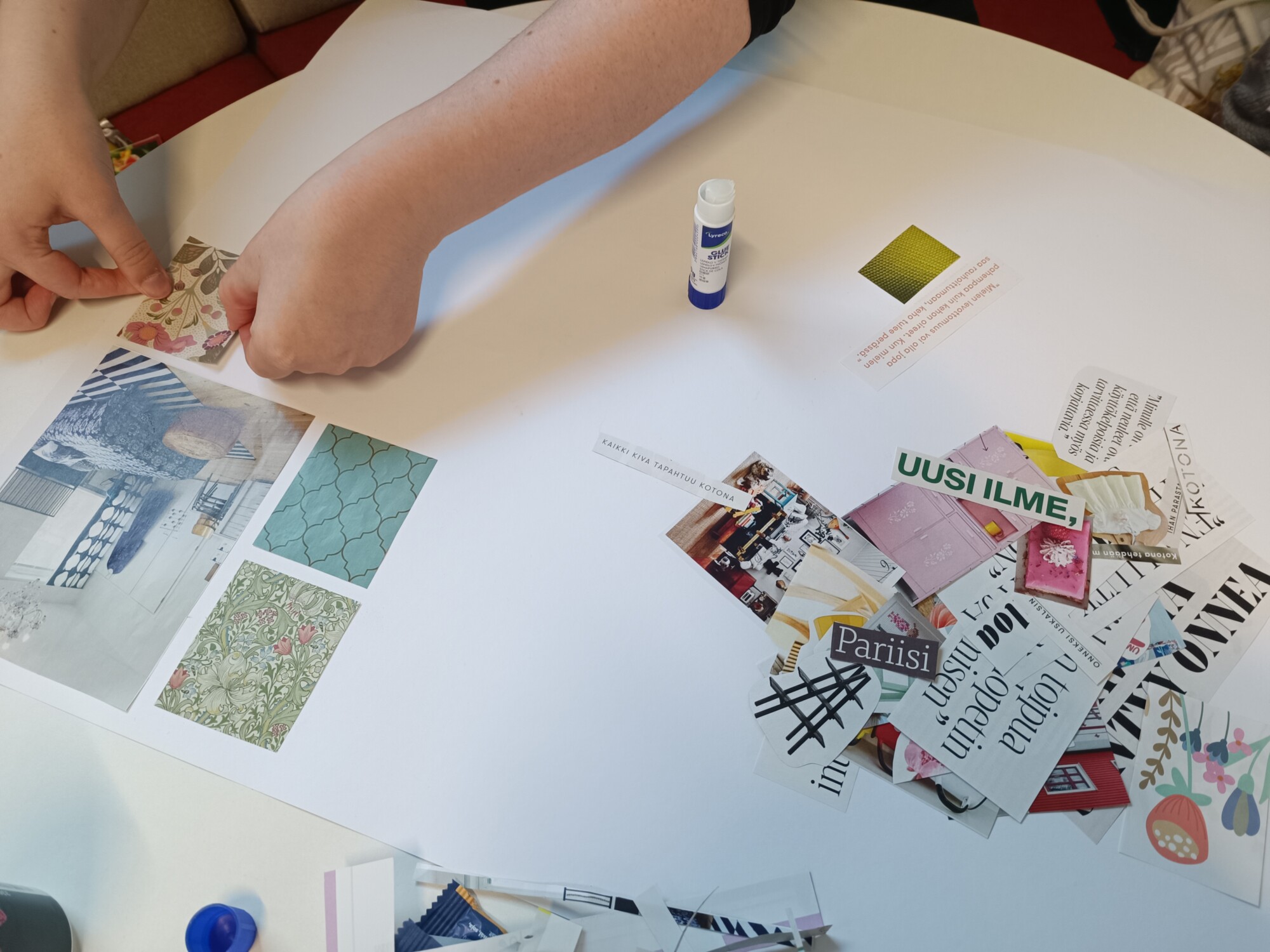 Henkilö järjestelee lehtileikkeitä ja kuvioituja kuvia valkoiselle paperille, liimapuikko käden ulottuvilla, ja luo kollaasia. Pöydällä on hajallaan erilaisia leikattuja sanoja ja kuvia.