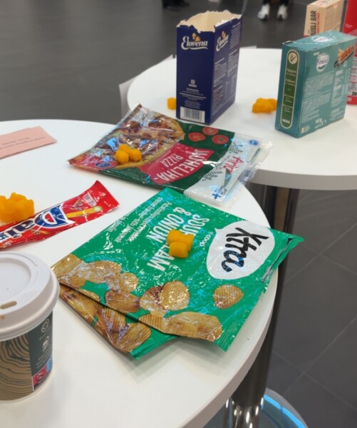 Kaksi pyöreää pöytää, joiden päällä on välipalapakkauksia, paperinen kahvikuppi, lattialle pudonneita juustovälipaloja ja avattuja pakkauksia. Taustalla näkyy tumma lattia ja epäselviä esineitä, mikä viittaa rentoon tai taukohetkeen.