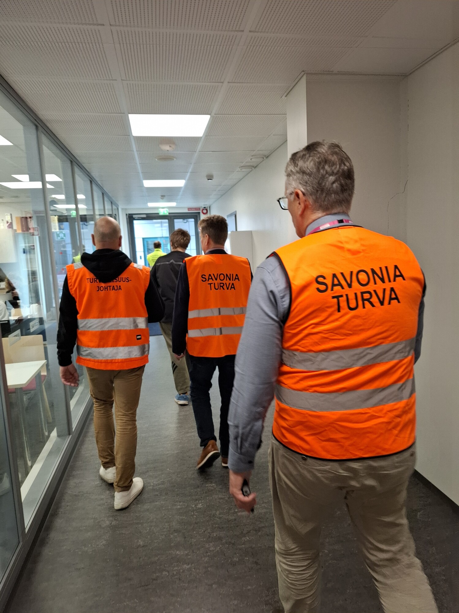 Neljä henkilöä, joilla on yllään oranssit heijastinliivit, joissa lukee ”SAVONIA TURVA”, kävelee lasiseinäistä ja valkoisilla kattolaatoilla varustettua käytävää pitkin. Kuva on otettu takaa, kun he etenevät eteenpäin.