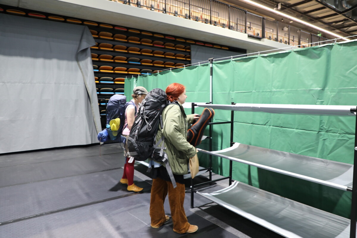 Kaksi henkilöä, joilla on selkäreput ja makuutarvikkeet, seisoo tyhjien kerrossänkyjen vieressä liikuntasalissa, jonka taustalla näkyy vihreitä väliseiniä ja pinottuja tuoleja.