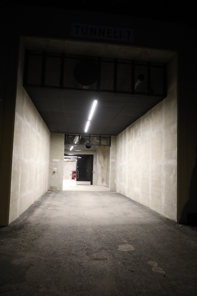 TUNNELI 7 -merkitty betonitunneli, jonka katossa palaa valo, johtaa kirkkaasti valaistuun avoimeen sisätilaan, jossa näkyy ovia ja putkia.