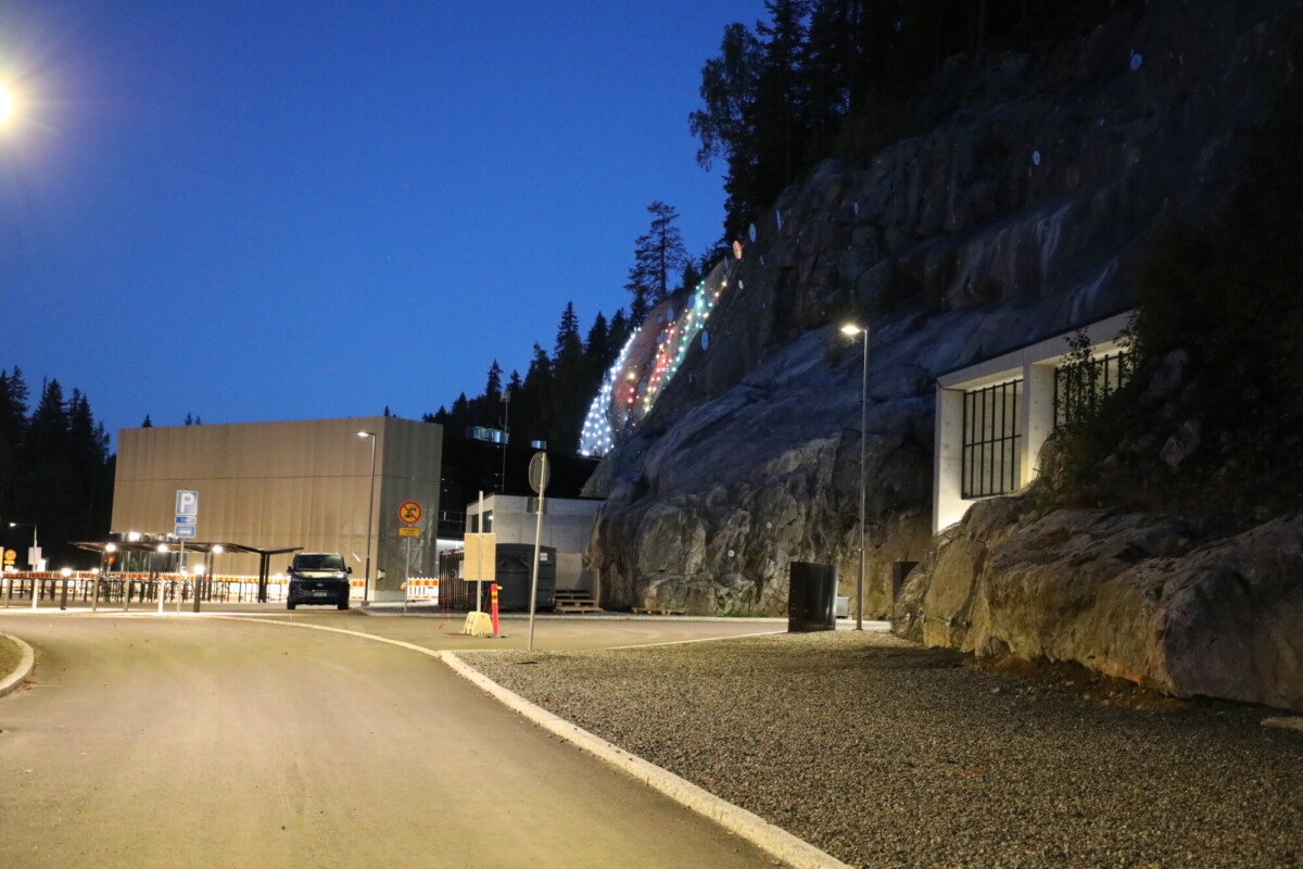 Hämärässä tie mutkittelee modernin rakennuksen ja kallioisen rinteen ohi; tarkastuspisteen vieressä on pysäköityjä autoja, ja rinteellä puiden seassa on ripustettu juhlavia valoja.