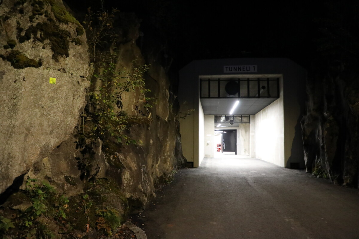 Yöllä kallioiden välissä näkyy hämärästi valaistu tunnelin sisäänkäynti, jossa lukee ”Tunneli 7”. Tunnelin sisällä on kirkas valaistus, mikä muodostaa jyrkän kontrastin pimeän ulkoilureitin kanssa.