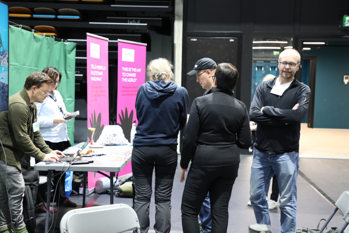 Sisätiloissa järjestetyssä tapahtumassa ihmiset ovat kokoontuneet pöydän ympärille kannettavien tietokoneiden ja papereiden kanssa. Paikalla näkyy värikkäitä banderolleja ja tuoleja, ja osa ihmisistä keskustelee, kun taas toiset työskentelevät tai seuraavat tilannetta.