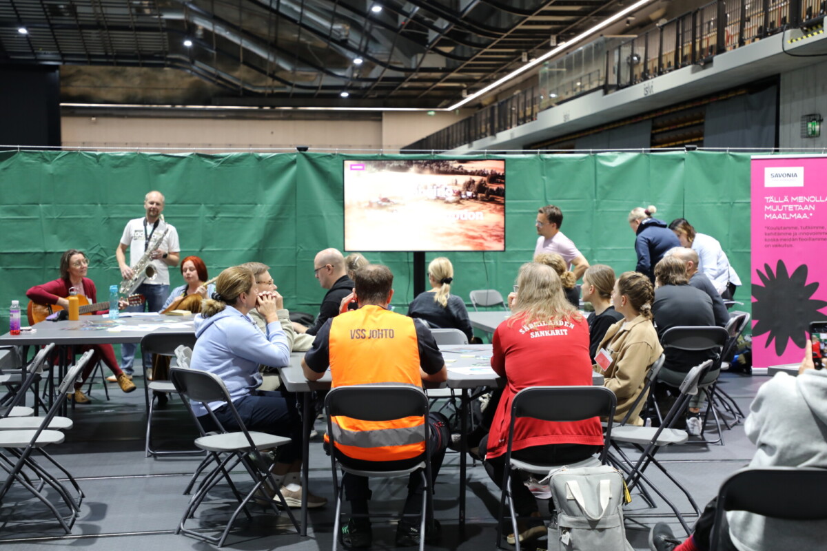 Ryhmä ihmisiä, jotka istuvat ja seisovat pöytien ympärillä suuressa sisätilassa; osalla on yllään univormut tai liivit, ja taustalla näkyy esitysnäyttö sekä vihreä väliseinä.