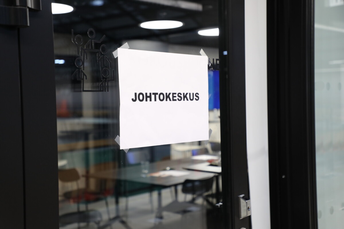 Lasiseen oveen on teipattu valkoinen paperikyltti, johon on painettu mustalla teksti ”JOHTOKESKUS”. Taustalla näkyy toimisto ja tyhjät tuolit.