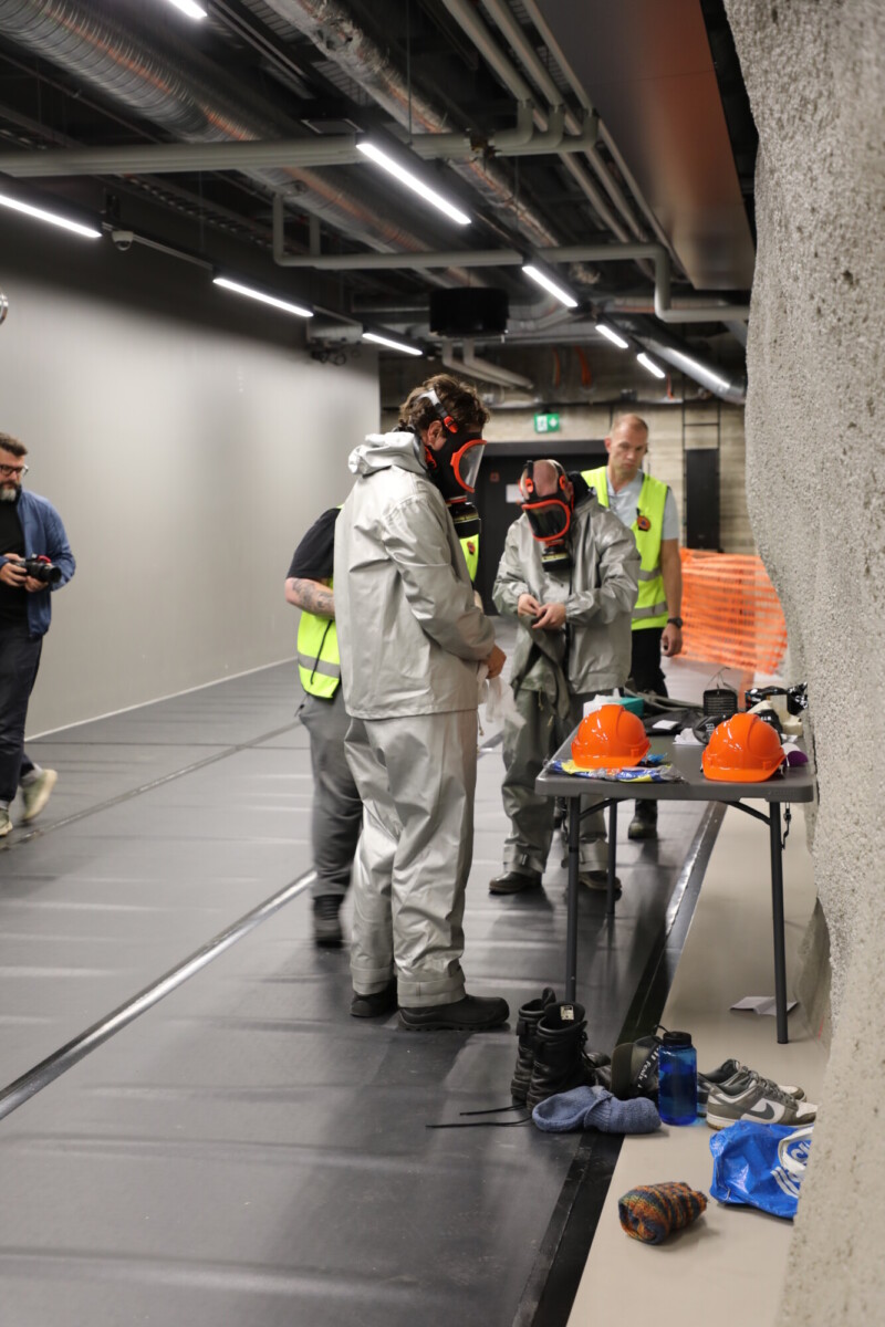 Useat hopeanvärisiä suojapukuja ja naamioita käyttävät henkilöt seisovat teollisuusrakennuksen sisätiloissa pöydän ääressä, jonka päällä on oransseja kypäröitä ja varusteita, ja muut seuraavat tilannetta lähistöllä.