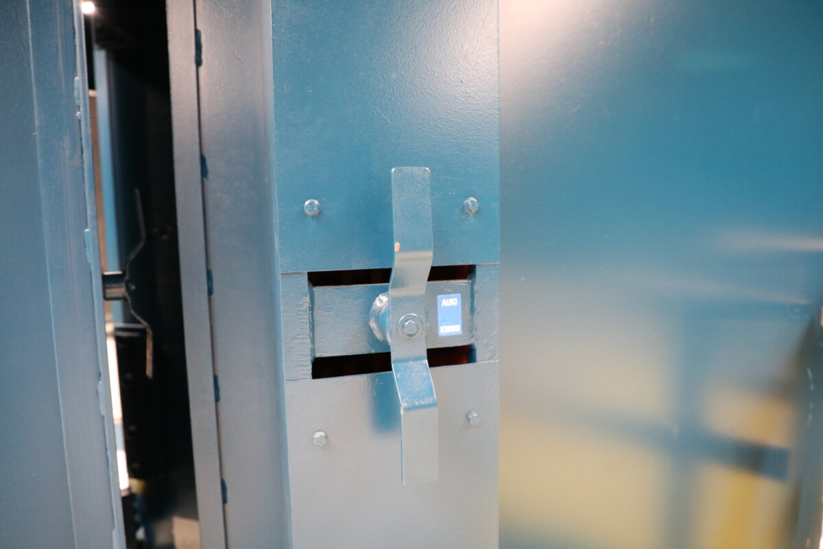 Lähikuva metallimekanismista, jossa on salpa ja pieni sininen painike, jossa lukee ”PUSH TO OPEN”, teräspinnalla; todennäköisesti osa turvaovea tai kassakaappia.