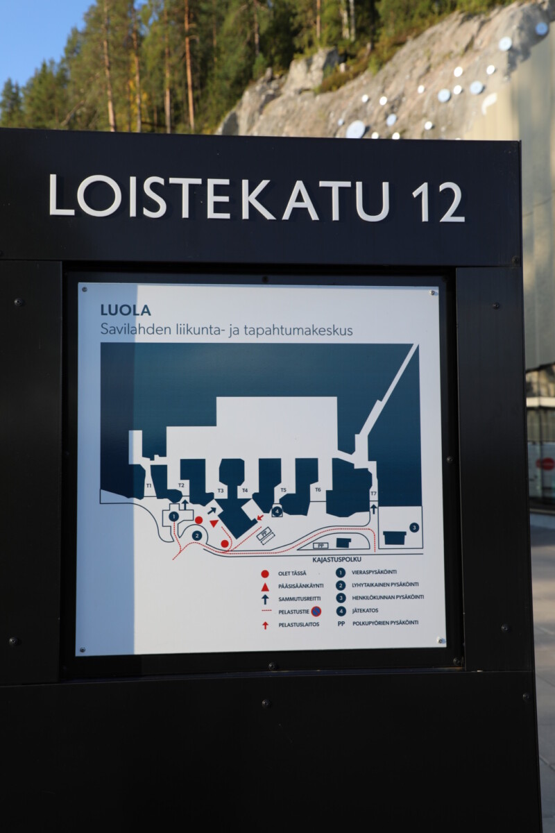 LOISTEKATU 12 -merkinnällä varustetussa karttakyltissä esitetään Savilahtin urheilu- ja tapahtumakeskuksen pohjapiirros suomeksi; siinä näkyvät rakennusten ääriviivat sekä pysäköintipaikkoja, pääsisäänkäyntiä ja infopistettä kuvaavat symbolit.
