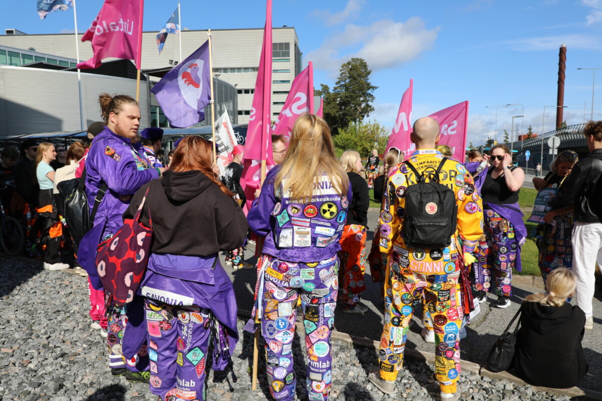 Ryhmä ihmisiä, joilla on yllään värikkäitä, lappuilla koristeltuja haalareita, seisoo yhdessä ulkona ja pitelee vaaleanpunaisia lippuja. Taustalla näkyy rakennuksia, puita ja muita ihmisiä sinisen taivaan alla.