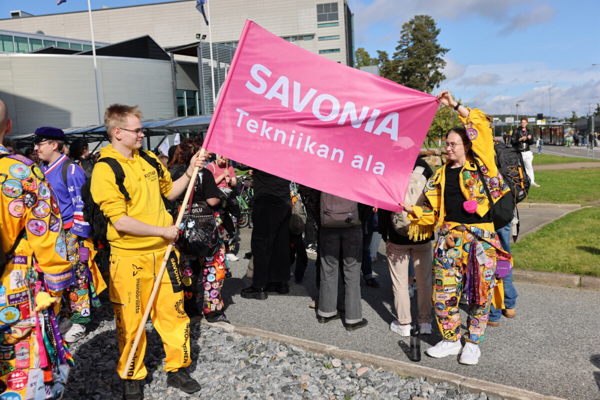 Kaksi värikkäissä haalareissa olevaa henkilöä pitelee ulkoilmatilaisuudessa vaaleanpunaista lippua, jossa lukee ”SAVONIA Tekniikan ala”. Heidän vieressään seisoo muita ihmisiä, ja taustalla näkyy moderni rakennus.