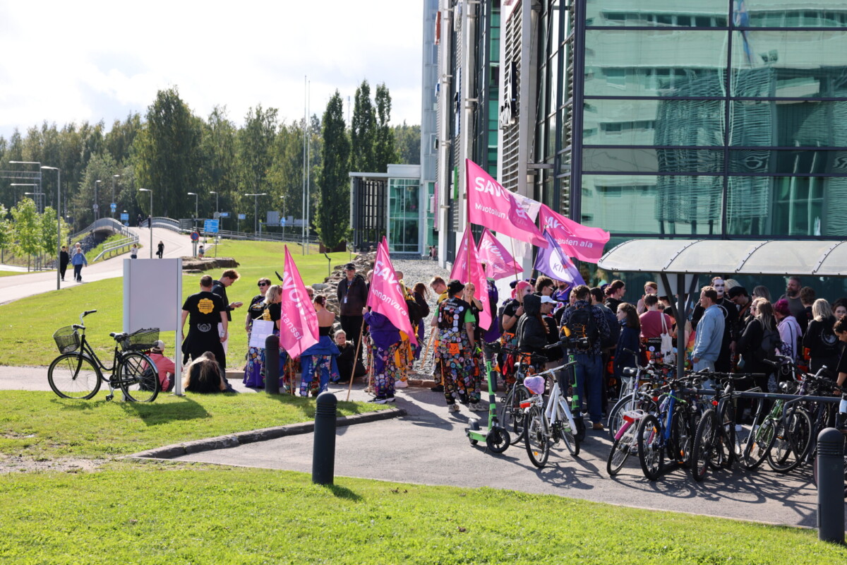 Ryhmä värikkäisiin vaatteisiin pukeutuneita ihmisiä, joilla on vaaleanpunaiset liput, on kokoontunut modernin lasirakennuksen eteen. Lähellä on pysäköitynä useita polkupyöriä, ja osa ihmisistä näyttää valmistautuvan mielenosoitukseen tai tapahtumaan. Taustalla näkyy puita ja nurmikkoa.