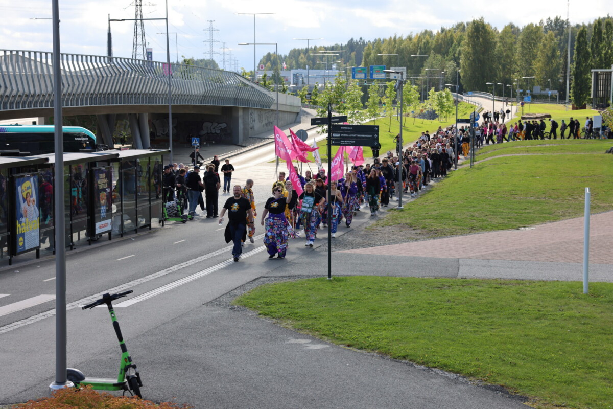 Suuri joukko ihmisiä marssii kaupungin jalkakäytävällä, ja osa heistä kantaa vaaleanpunaisia lippuja. Kuvassa näkyy bussipysäkki, skoottereita, vehreyttä ja läheisiä teitä osittain pilvisen taivaan alla.