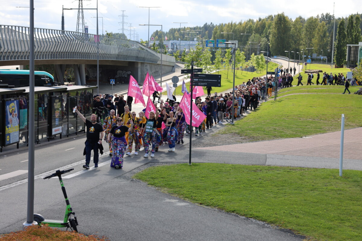 Suuri joukko ihmisiä, joista osa pitää kädessään vaaleanpunaisia lippuja, marssii aurinkoisena päivänä jalkakäytävää pitkin bussipysäkin ja vihreän nurmikon vieressä. Lähellä on pysäköitynä vihreä skootteri. Taustalla näkyy puita ja katukyltejä.