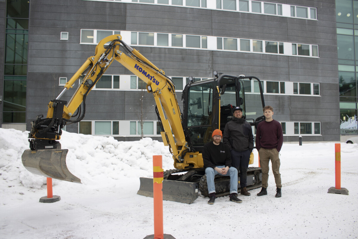 Kolme henkilöä seisoo keltaisen Komatsu-kaivinkoneen edessä lumisella maalla, lähellä toimistorakennusta, jossa on paljon ikkunoita. Etualalla näkyy oransseja pylväitä.
